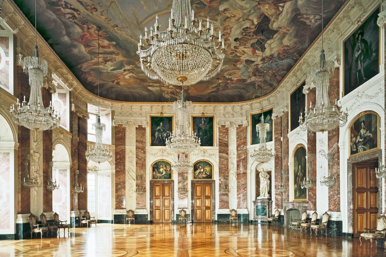 Foto des Rittersaals im Schloss Mannheim. Er hat reich dekorierte Wände, ein Deckengemälde und große Deckenleuchter.
