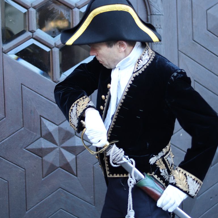 Foto eines Mannes in einer Uniform aus dem 19. Jahrhundert. Er steht vor einer Holztür und will das dahinter befindliche Gebäude betreten. Er ist gerade dabei, seinen Säbel zu ziehen.