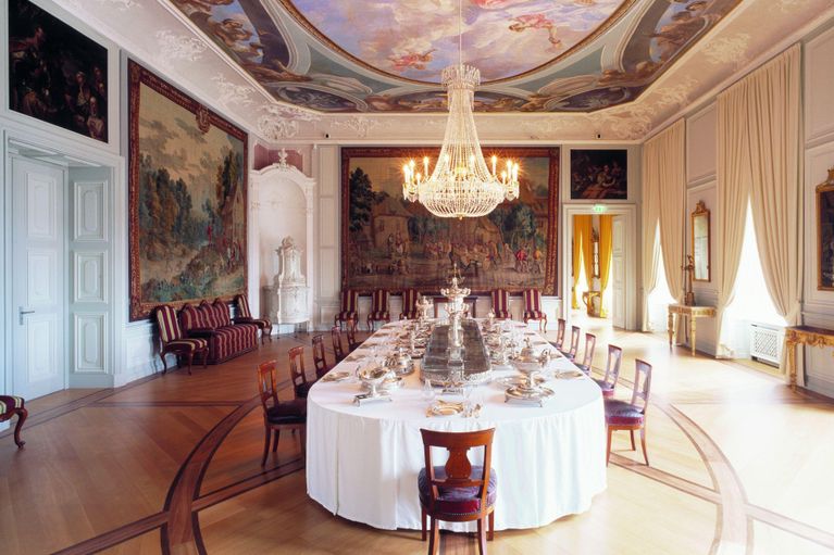 Foto eines Saals im Schloss Mannheim. In der Mitte steht eine mit silbernem Geschirr gedeckte Tafel. An den Wänden hängen kostbare Teppiche und Vorhänge