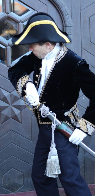Foto eines Mannes in einer Uniform aus dem 19. Jahrhundert. Er steht vor einer Holztür und will das dahinter befindliche Gebäude betreten. Er ist gerade dabei, seinen Säbel zu ziehen.